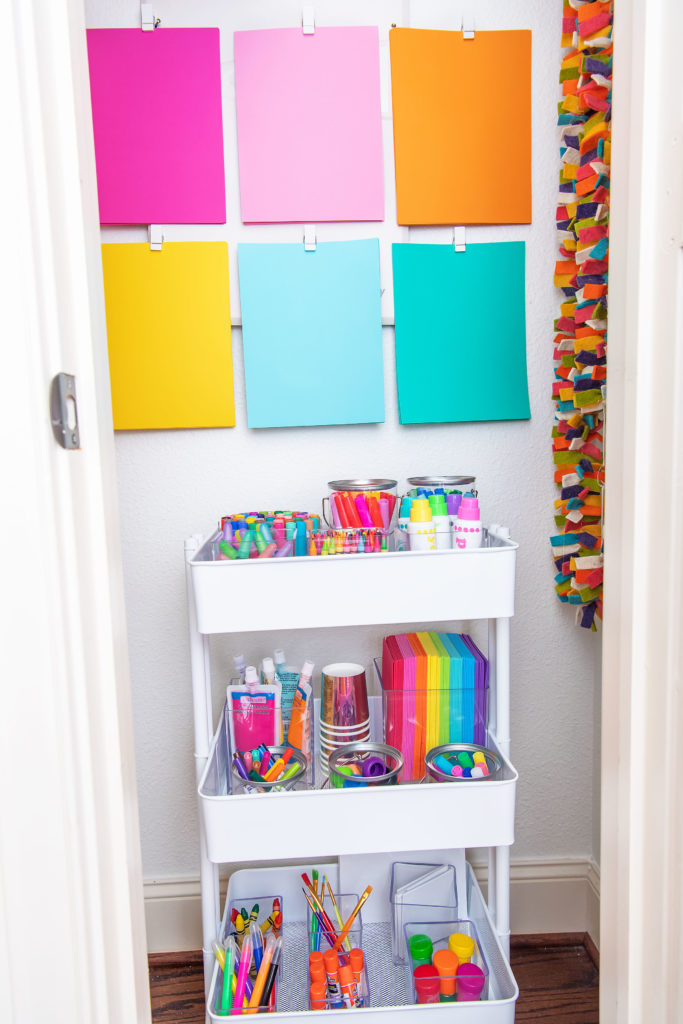 Craft Closet by popular Houston lifestyle blog, Cake and Confetti: image of a craft closet filled with paper scissors, paint brushes, cardstock paper, bakers twin, tape, felt garland, pom pom balls, and a metal card on wheels stocked up with acrylic containers filled with markers, crayons, dot markers, paper, glue sticks, and glitter glue. 