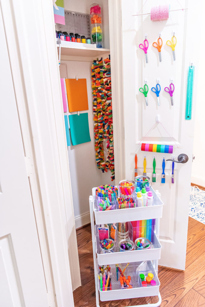 Craft Closet by popular Houston lifestyle blog, Cake and Confetti: image of a craft closet filled with paper scissors, paint brushes, cardstock paper, bakers twin, tape, felt garland, pom pom balls, and a metal card on wheels stocked up with acrylic containers filled with markers, crayons, dot markers, paper, glue sticks, and glitter glue. 