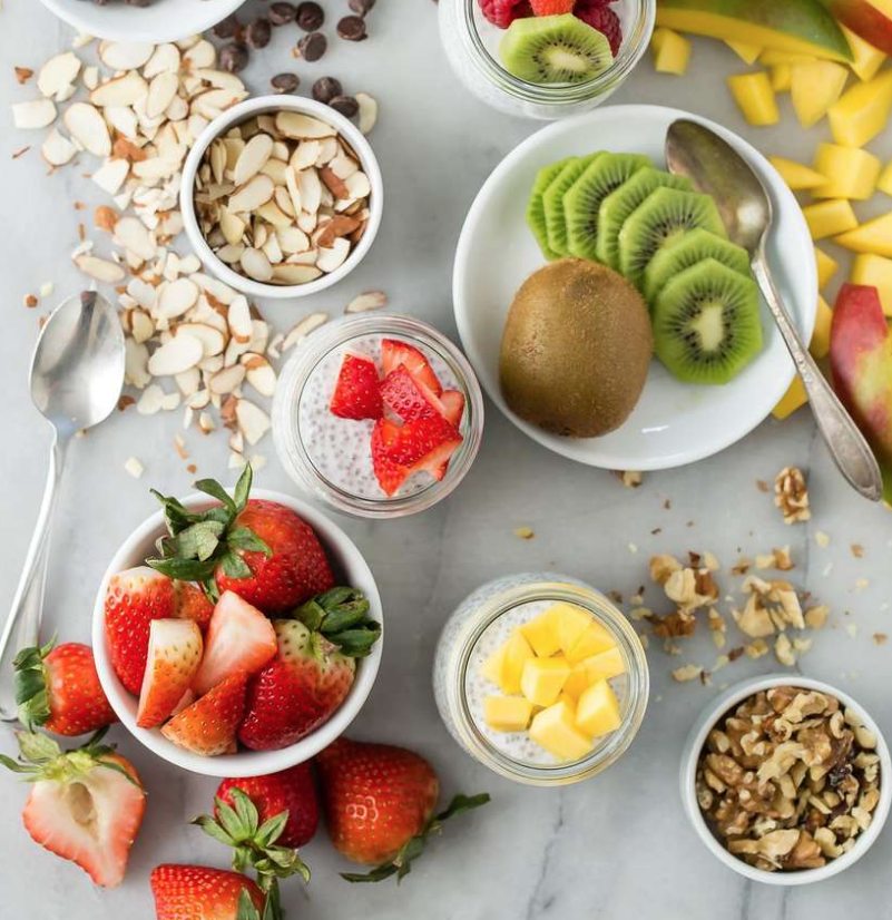 Easy Memorial Day Brunch Recipes by poplar Houston lifestyle blog, Cake and Confetti: image of coconut chia pudding in mason jars next to a bowl of strawberries, a bowl of sliced almonds, a bowl of walnuts, and a bowl of sliced kiwi. 